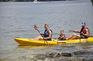 A woman, an man, and a child wearing life jackets paddle a large kayak on a lake near Sandy Island.