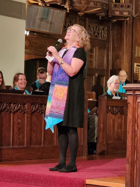A blonde woman wearing glasses, a black dress, and a rainbow ministerial stole holds a microphone in both hands as she looks up and smiles in the front of the sanctuary