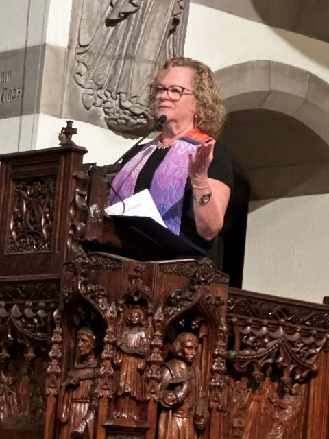 A blonde woman wearing glasses, a black dress, and a rainbow ministerial stole stands in a large carved wooden pulpit with her hands outstretched