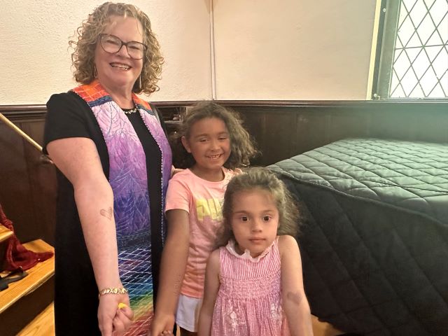 A blonde woman wearing glasses, a black dress, and a rainbow ministerial stole stands with two children in pink dresses by the piano in the Parish Hall
