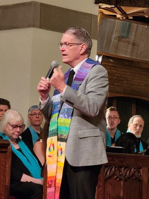 A gray-haired man wearing glasses, a gray suit jacket, black pants, and a rainbow ministerial stole stands holding a microphone and addressing the congregation