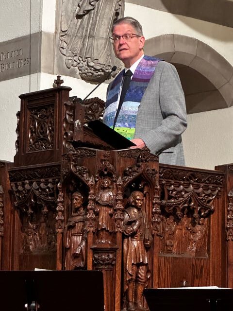 A gray-haired man wearing glasses, a gray suit jacket, a white shirt, a black tie, and a rainbow ministerial stole speaks from a large carved wooden pulpit