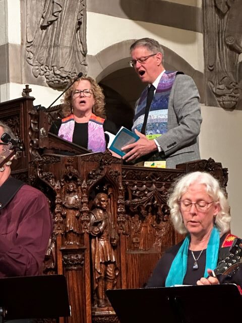A blonde woman wearing glasses, a black dress, and a ministerial stole and a gray-haired man wearing glasses, a gray suit jacket, and a rainbow ministerial stole stand in a large wooden pulpit and sing along with the choir below