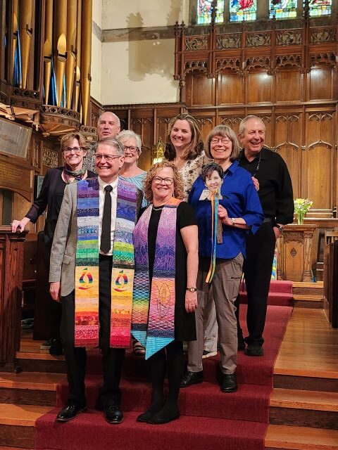 A group of eight adults, with a man and a woman wearing rainbow ministerial stoles in front, stand in the sanctuary of a church.