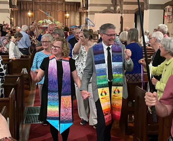 A smiling blonde woman wearing glasses, a black dress, and a ministerial stole and a smiling gray-haired man wearing glasses, a gray suit jacket, and a rainbow ministerial stole walk down the aisle of the sanctuary, followed and feted by congregants waving ribbons in various colors