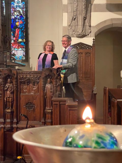 A blonde woman wearing glasses, a black dress, and a ministerial stole and a gray-haired man wearing glasses, a gray suit jacket, and a rainbow ministerial stole stand in a large wooden pulpit with a burning chalice in the foreground