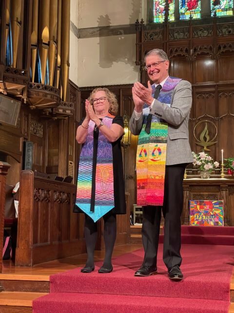 A blonde woman wearing glasses, a black dress, and a ministerial stole and a gray-haired man wearing glasses, a gray suit jacket, and a rainbow ministerial stand before the chancel and clap